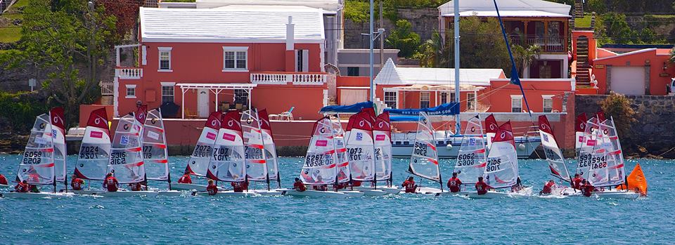 A group of small sailboats with red and white sails racing on blue water, with several pink and white buildings visible along the shoreline in the background.