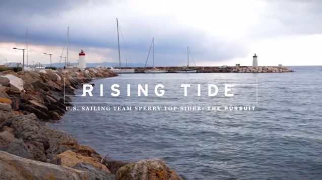 Rocky shoreline with calm water, a lighthouse, and several sailboat masts in the distance under a cloudy sky. Text on the image reads: 'Rising Tide. U.S. Sailing Team Sperry Top-Sider, The Pursuit.
