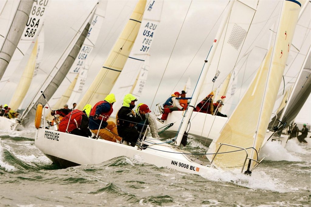 Several sailboats race closely together on choppy water, with crews wearing bright jackets and hats. The scene is dynamic, with angled sails and splashing waves under a cloudy sky.