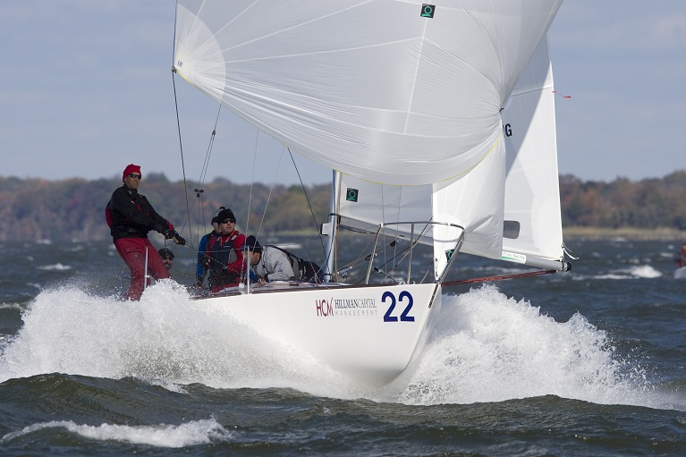 A white sailboat with the number 22 sails through choppy waters, creating splashes. Four crew members in jackets and hats are on board. Trees and a cloudy sky are visible in the background.