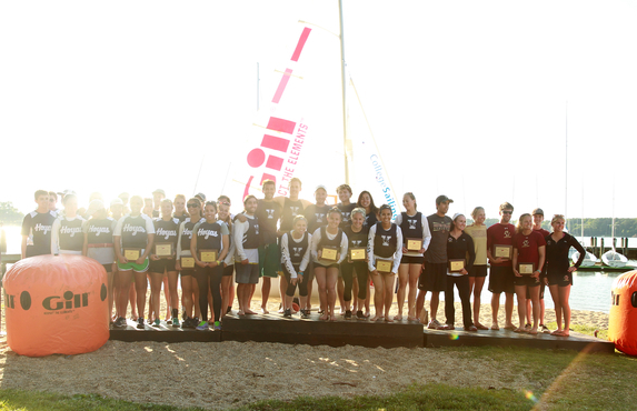 A group of young athletes stands together on a podium outdoors, holding plaques and medals, with sailboats and water in the background on a sunny day. Large orange buoys and event banners are visible.
