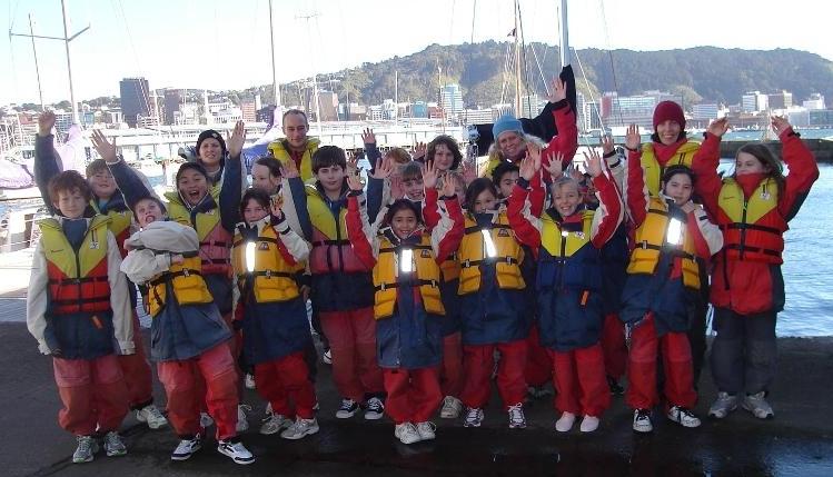 A group of children and a few adults wearing life jackets and outdoor clothing pose and smile with raised arms by a marina, with boats and a cityscape visible in the background.