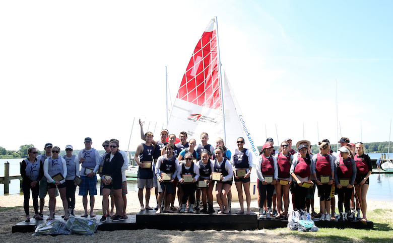 A group of people stand on and around a podium outdoors near a lake, holding awards. Behind them are sailboats and a large sail with red and white patterns. The group appears to be celebrating at a sailing event.