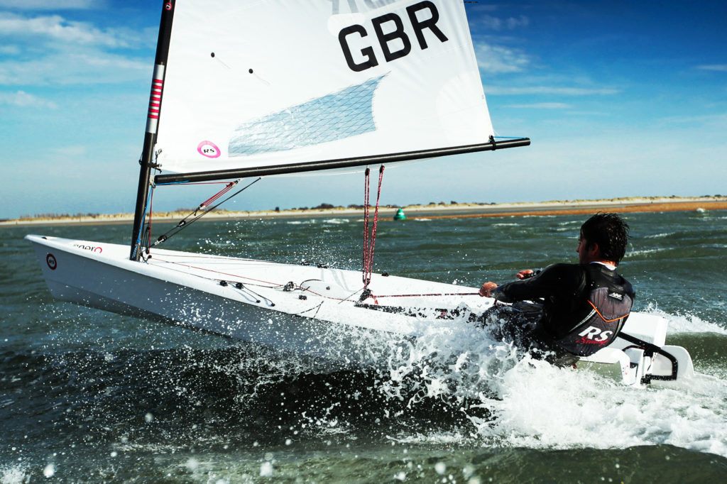 A person sails a small white dinghy labeled 'GBR' through choppy waters, wearing a wetsuit and life jacket. Spray splashes around the boat, and a sandy coastline is visible in the background under a blue sky.