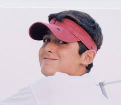 A young person with short dark hair wearing a red visor and a white shirt looks over their shoulder and smiles.