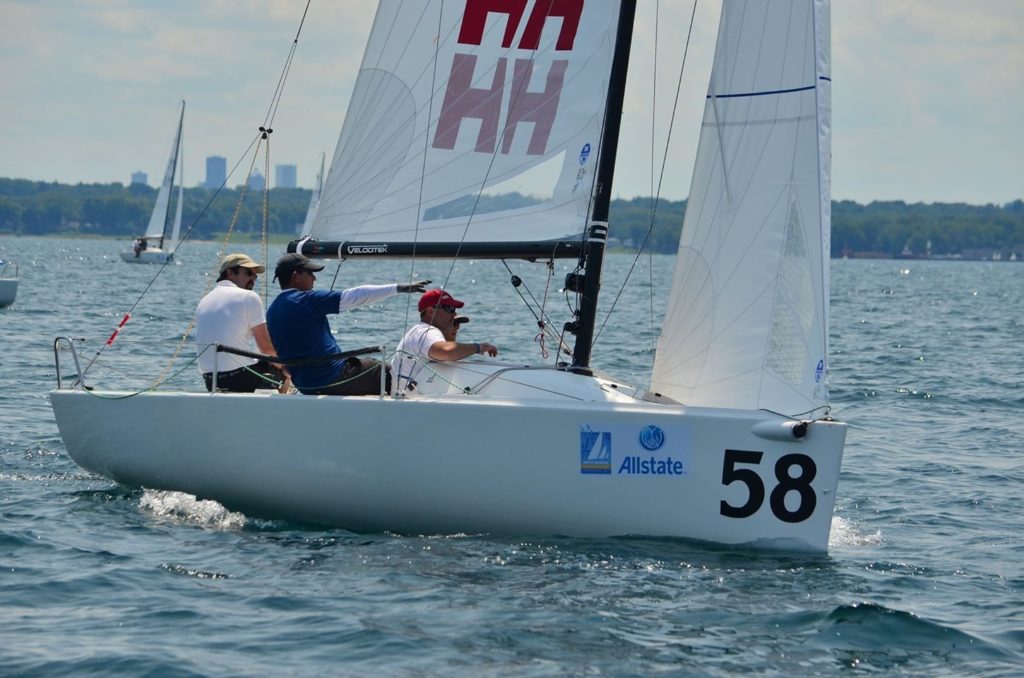 Three people sail a white racing boat numbered 58, with Allstate branding, on a sunny day. One person points ahead while others steer. Trees and distant buildings are visible on the shore under a partly cloudy sky.