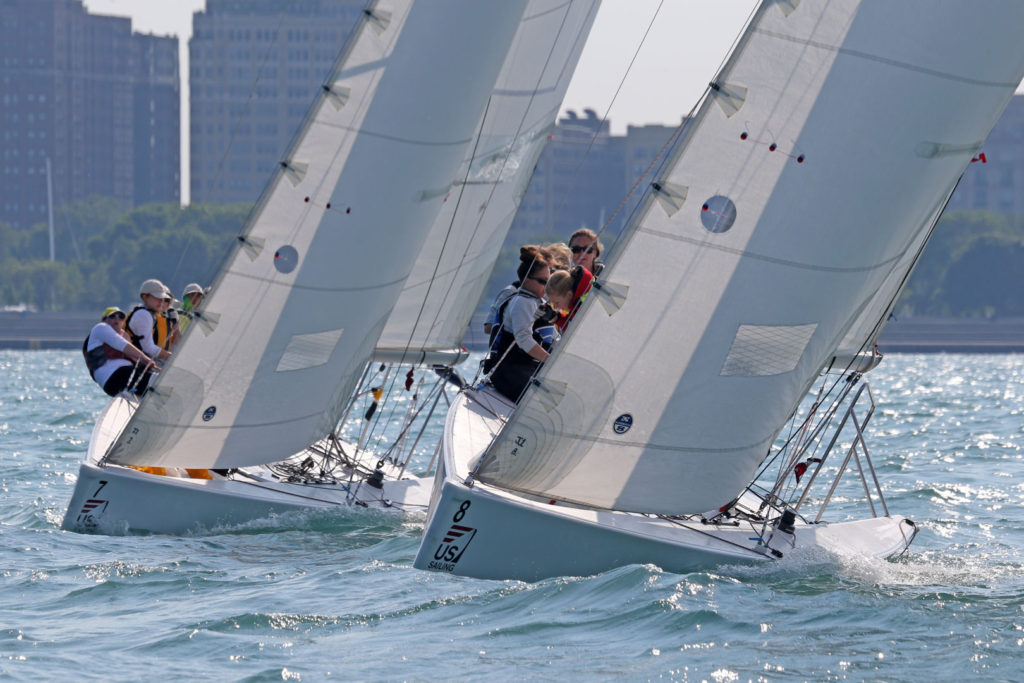 Two sailboats with crews racing on a sunny day, leaning into the wind with city buildings and trees visible in the background.