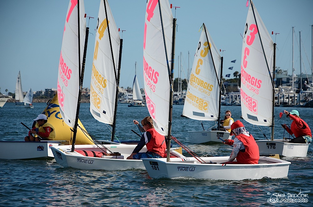 Children in small sailboats wearing life jackets participate in a sailing race, maneuvering around a yellow buoy on a sunny day with other boats and a marina visible in the background.