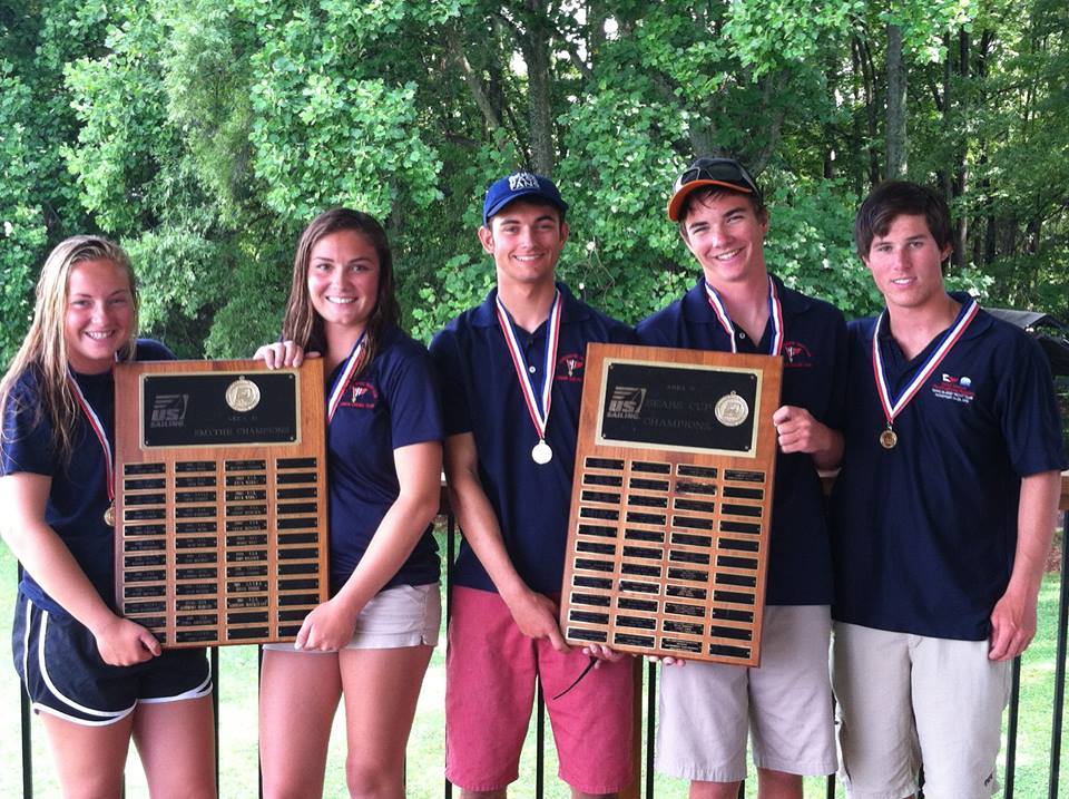 Five smiling young adults wearing medals and navy shirts stand outdoors, holding two large award plaques. Trees fill the background, and the group appears happy and celebratory.