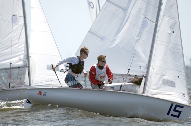 Two sailors wearing life vests and sunglasses compete in a small sailboat with the number 15, navigating choppy waters on a sunny day during a sailing race. White sails and other boats are visible in the background.