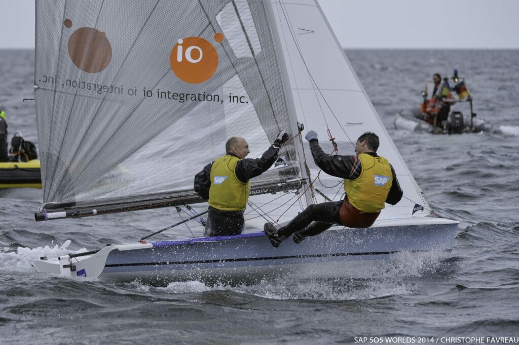 Two sailors in yellow vests race a small sailboat on choppy waters, leaning out to balance. The sail displays a large orange dot and the text 'io integration, inc.' Support boats follow in the background.
