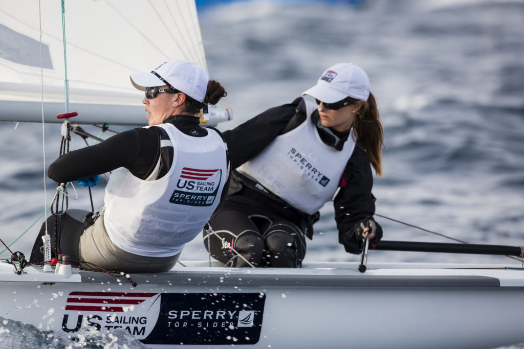 Two women sailors wearing 'US Sailing Team' vests and white hats compete in a small sailboat on choppy water, focused and handling ropes and equipment during a race.
