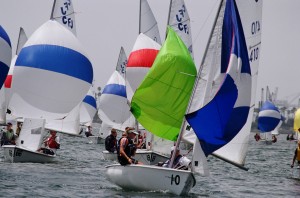 Several small sailboats with colorful spinnaker sails, including green, blue, red, and yellow, compete closely together on the water during a sailing race. Sailors are actively maneuvering their boats.