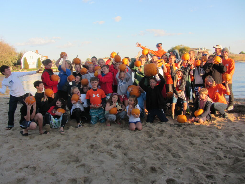 A large group of children and adults stand on a sandy beach holding small pumpkins and smiling. The background shows water, houses, and a clear sky on a sunny day.