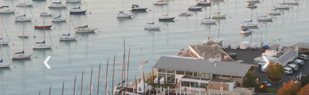 A marina filled with numerous sailboats on calm water, with a building and parked cars along the shore in the foreground. Two white arrow icons are visible on each side of the image.