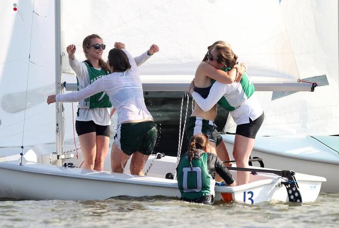 Four young women celebrate joyfully on a small sailboat, with two hugging and two raising their arms in excitement. Another person in a life vest stands in the shallow water beside the boat.