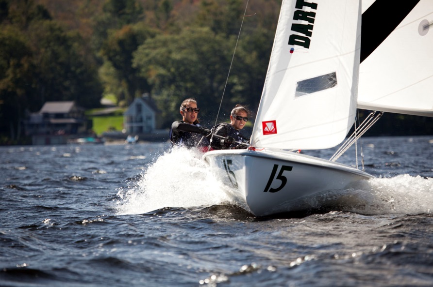 Two people wearing sunglasses and life vests sail a small white boat with the number 15 on a lake, water splashing around them, with houses and trees visible in the background.