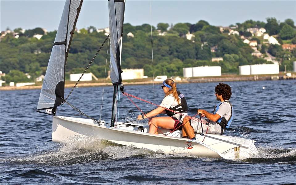 Two people wearing life jackets sail a small white sailboat on a body of water, with green hills and white buildings visible in the background under a clear sky.