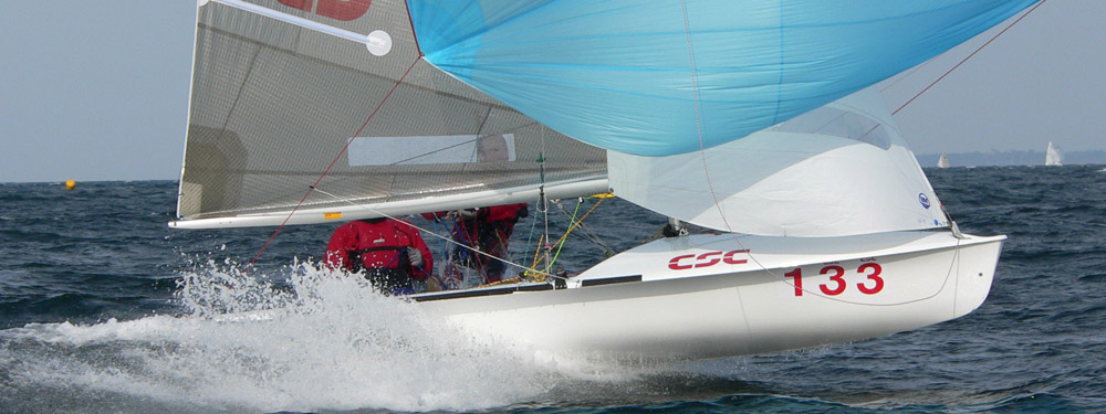 A white sailboat with the number 133 and blue and white sails glides quickly over choppy water, creating a spray. Two people in red jackets are visible on the boat.