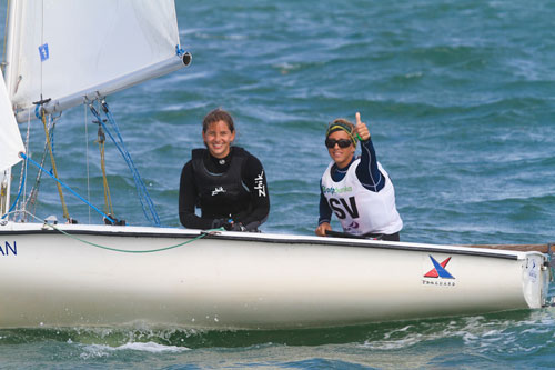 Two sailors in a small sailboat on the water, both smiling. One sailor is giving a thumbs-up while the other holds the tiller. Both are wearing wetsuits and life jackets. The sea appears calm and the sun is shining.