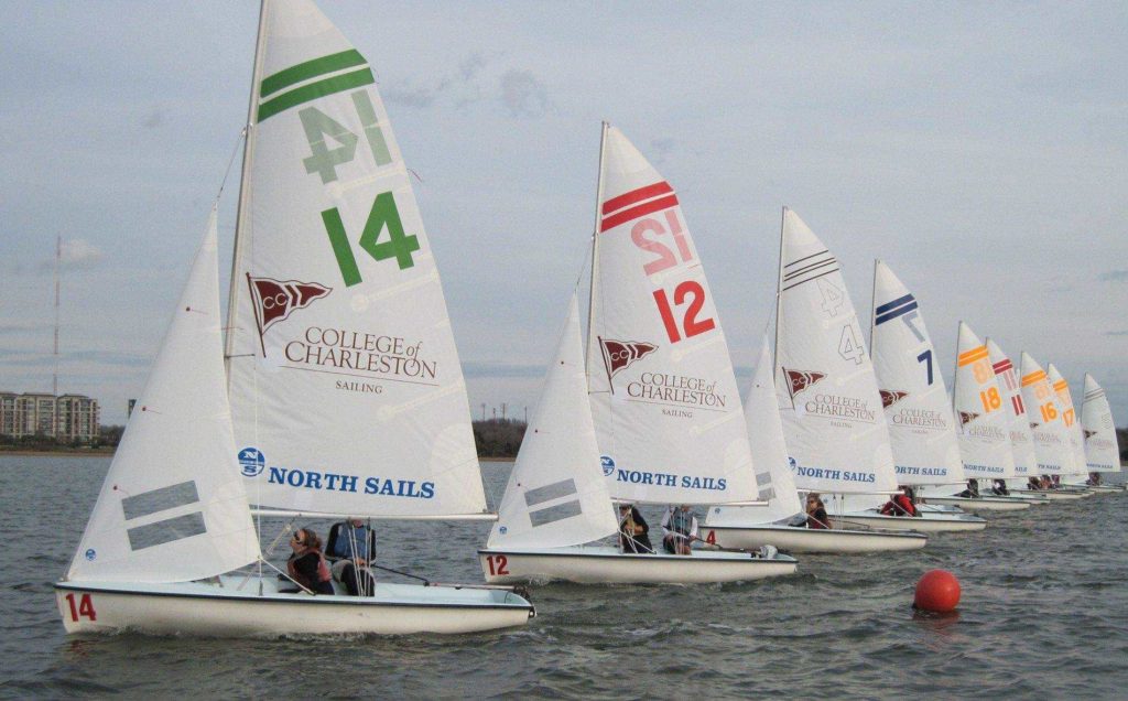 A row of small sailboats from the College of Charleston sailing team, each with colored numbers and sails, race closely together on the water near a shoreline.