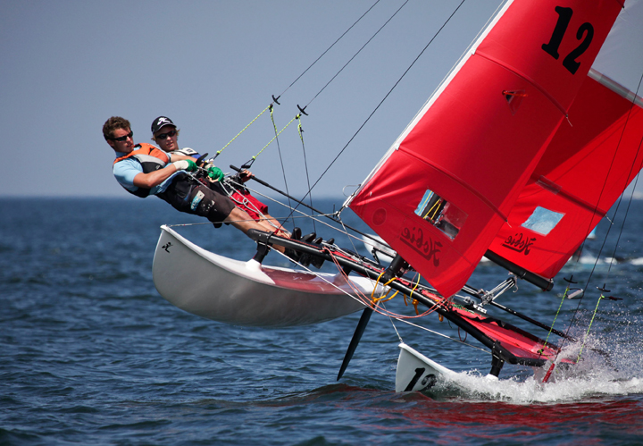 Two people sailing a catamaran with bright red sails, leaning out over the water as the boat tilts, creating a dynamic action scene on a sunny day.
