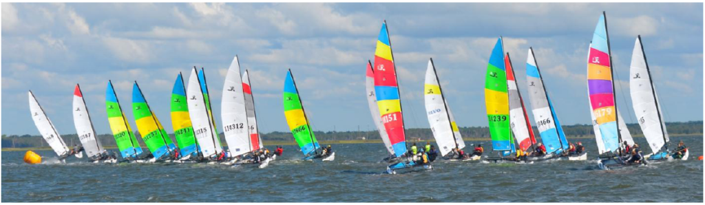 A group of colorful sailboats with vibrant sails race closely together on a choppy body of water under a cloudy sky, with a yellow buoy visible on the left side of the image.