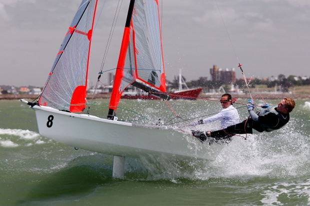 Two sailors steer a small sailboat numbered 8, leaning out over the water as waves splash against the hull. The boat has bright red and gray sails, and buildings are visible in the background.