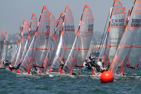 Several sailboats with transparent sails and red trim race closely together on the water, with sailors focusing intently. A bright red buoy floats in the foreground, and city buildings are visible in the background.