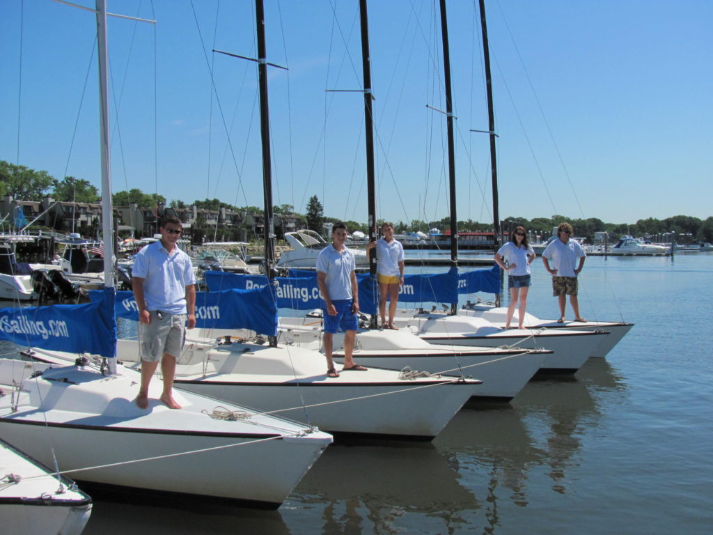 Five people stand on the bows of five white sailboats docked side by side on a sunny day, with blue skies, calm water, and buildings and boats in the background. Blue banners with “BeSailing.com” are visible on the boats.