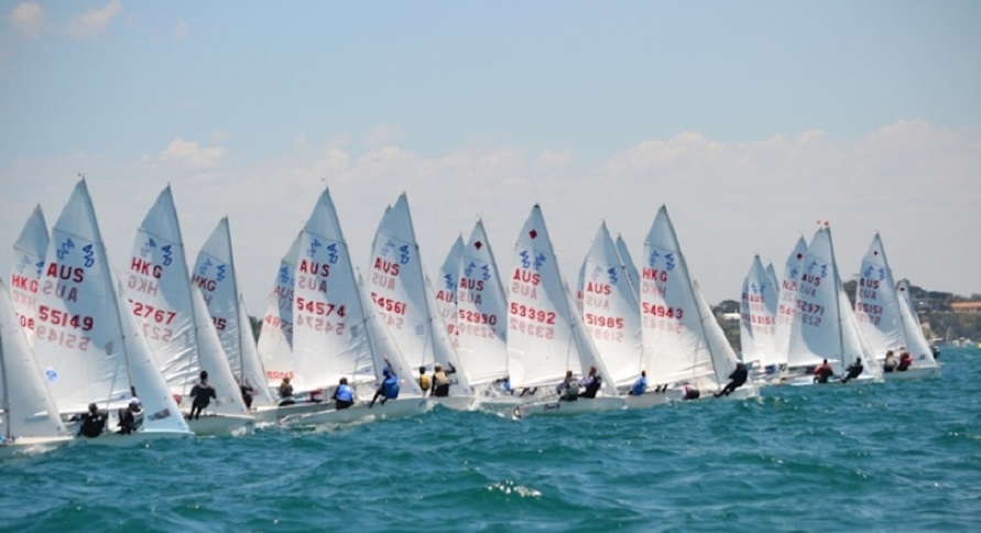 A row of sailboats with white sails, each marked with numbers and country codes, compete in a sailing race on blue water under a clear sky. Sailors are visible on each boat, leaning to balance.