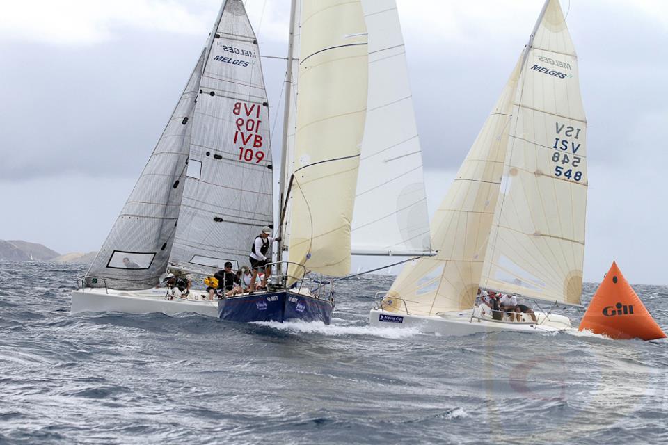 Three sailboats race closely around an orange buoy on choppy water under a cloudy sky, with crew members actively adjusting sails and steering. The sails display various numbers and logos.