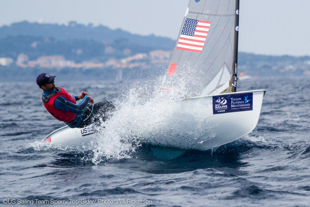 A sailor in a life jacket leans back on a small sailboat as it splashes through waves, with an American flag on the sail and coastal buildings in the blurred background.
