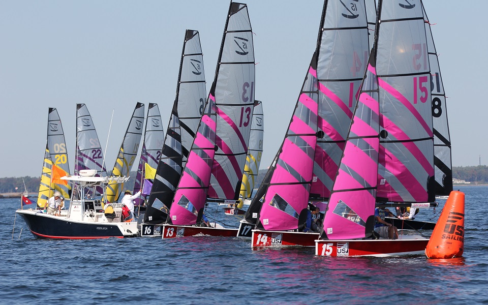 Several colorful sailboats with pink, yellow, and black sails race closely together near an orange buoy on the water, accompanied by a white motorboat, under a clear blue sky.