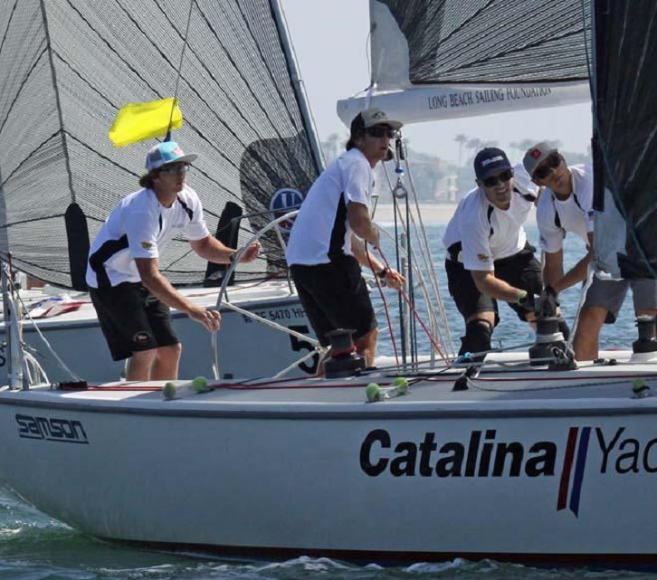 Four men in white shirts and dark shorts sail a Catalina yacht on a sunny day, with a yellow flag attached to a mast.