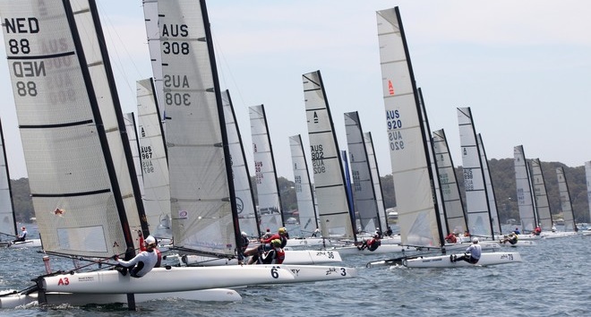 A group of sailboats with numbered sails and country codes compete in a race on a body of water, with sailors onboard and a hilly shoreline visible in the background.