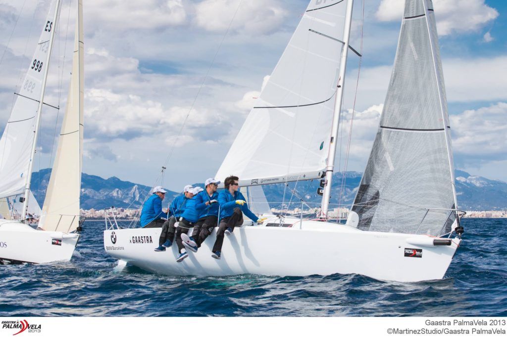 Four sailors in blue jackets sit on the edge of a white sailboat named Gaastra, racing on the ocean near a coastline with mountains and clouds in the background. Other sailboats are partially visible nearby.