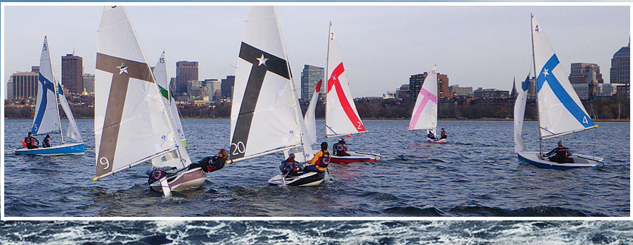 Sailboats with numbered sails race on a choppy river with several people aboard each, and a city skyline with tall buildings is visible in the background under a cloudy sky.
