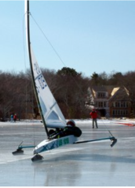 A person rides an iceboat with a sail across a frozen lake, passing by two people standing on the ice and houses in the background.