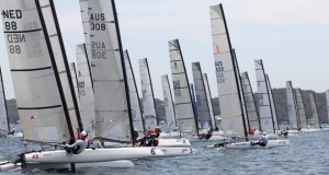 Several sailboats with tall sails race closely together on a body of water, with sailors visible onboard. Land with trees can be seen in the background under a cloudy sky.