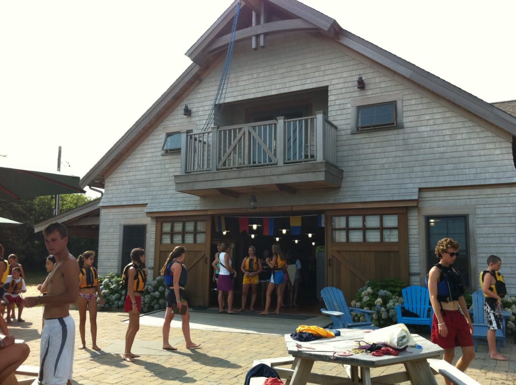 A group of people, many wearing life jackets, stand and chat outside a large wooden boathouse with a balcony. Blue chairs and tables are in the foreground on a sunny day.
