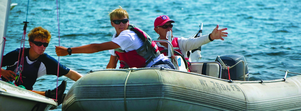 Three people on a small inflatable boat on the water, two young men wearing sunglasses and life jackets, and an older man pointing toward the distance, appearing to give instructions.
