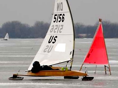 A person operates an iceboat with a white sail marked 'DN 5156 US' on a frozen lake, passing by a bright red triangular marker; trees are visible in the background.
