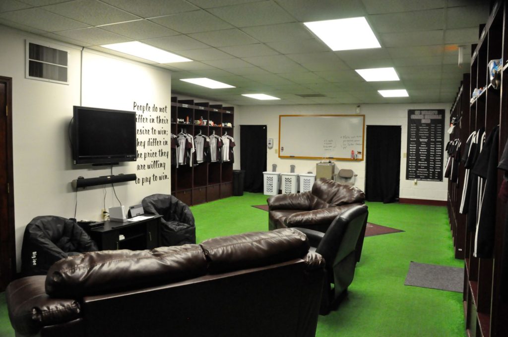 A lounge area in a locker room with leather couches, a TV on the wall, open lockers with jerseys, bean bags, and a whiteboard in the background. The floor is green carpet, resembling grass.
