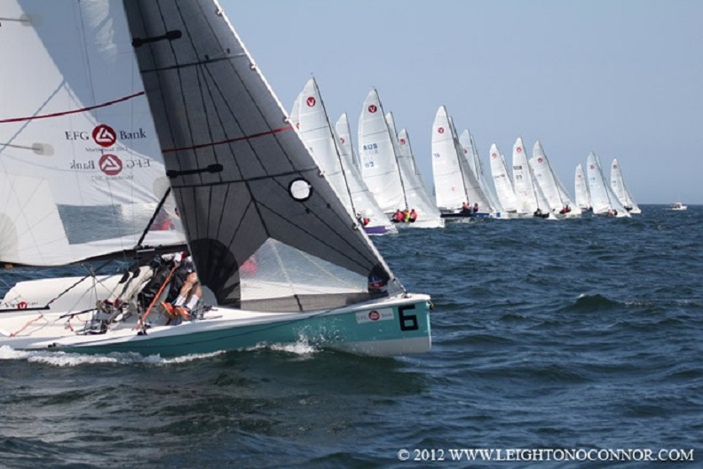 A fleet of small sailboats races on open water under a clear sky. The nearest boat has the number 6 and two visible sailors, while many others follow behind in the distance.