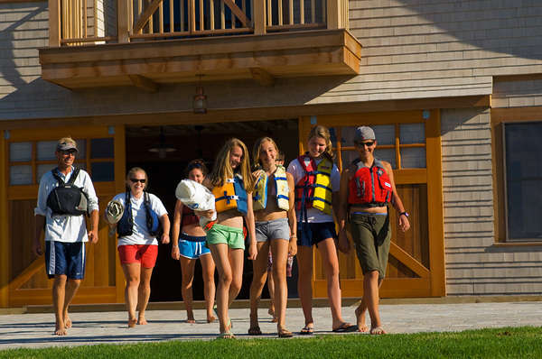A group of people wearing life jackets and casual summer clothes walk together outside a wooden building, smiling and enjoying a sunny day.