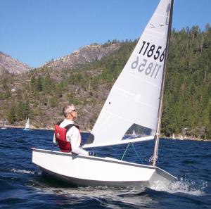 A person wearing a red life jacket sails a small white sailboat on a lake surrounded by pine-covered hills and rocky mountains under a clear sky.