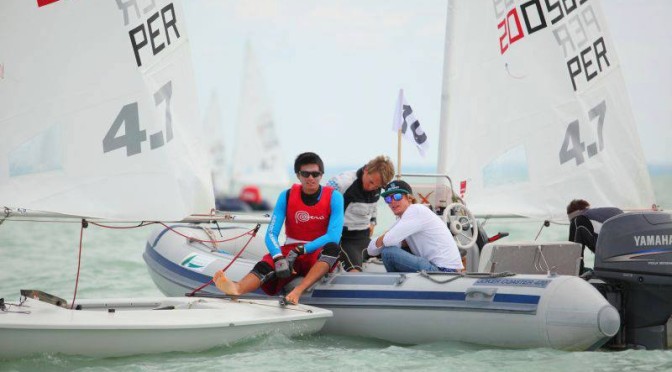 Three people sit in a motorboat next to a small sailboat on the water. Two wear sunglasses and one is holding onto the sailboat. Sailboats are visible in the background on a cloudy day.