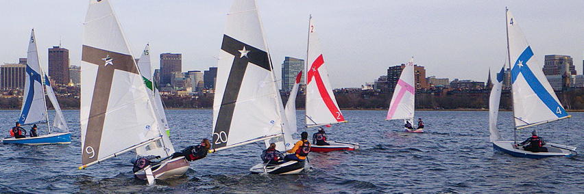 Several small sailboats with colorful sails race on a choppy river, with people on board. A city skyline with tall buildings is visible in the background under a cloudy sky.