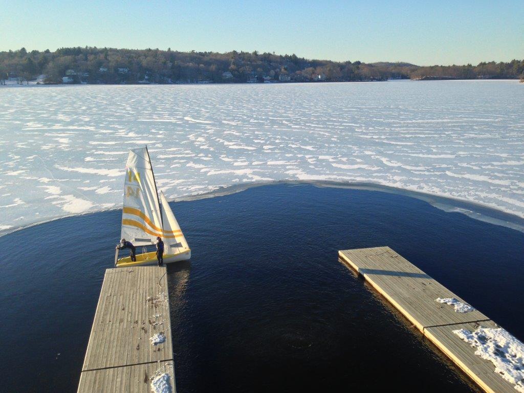 A small sailboat with two people is docked at a wooden pier, surrounded by a mostly frozen lake with a large circular area of open water near the docks. Snow covers the ice and distant trees line the shore.